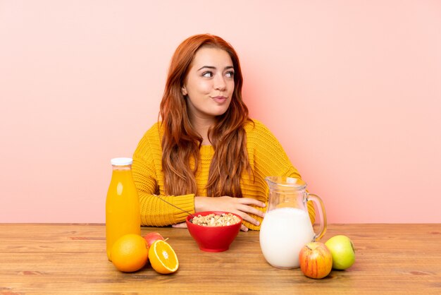 Ragazza rossa adolescente facendo colazione in un tavolo in piedi e guardando al lato