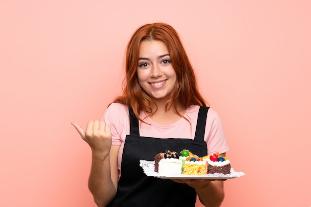 Ragazza rossa adolescente in possesso di un sacco di diverse mini torte su rosa isolato che punta verso il lato per presentare un prodotto