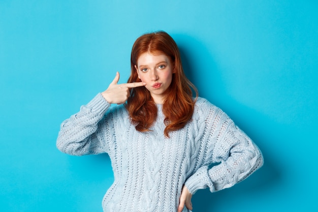 Ragazza rossa carina in maglione che colpisce la sua guancia, fissando la telecamera impertinente, in piedi su sfondo blu.