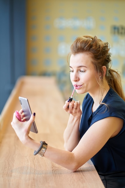 Ragazza rossa che mette il rossetto da sola, guardando alla riflessione su smartphone