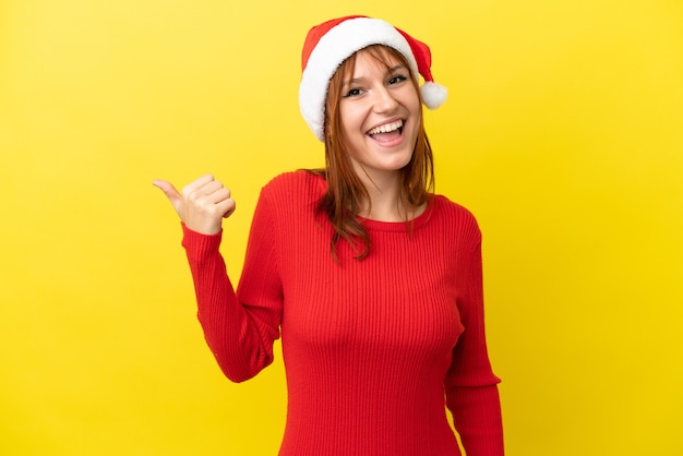 Ragazza rossa con cappello natalizio isolato su sfondo giallo che punta al lato per presentare un prodotto