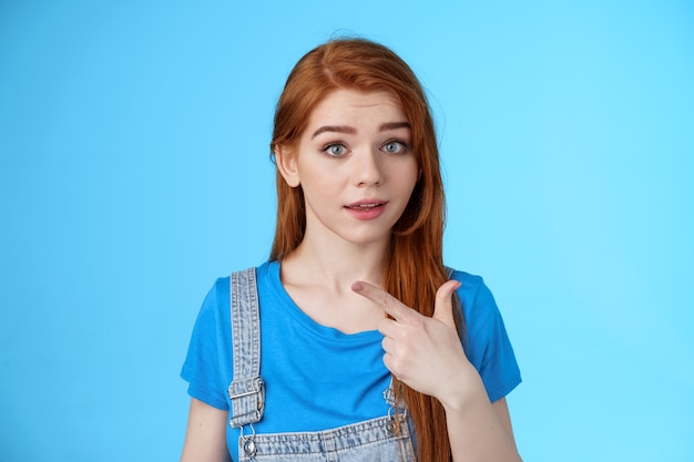 Ragazza rossa curiosa che punta a sinistra interrogata, incuriosita dall'interessante spazio di copia sembra domandata macchina fotografica imboscata, alza le sopracciglia confuse, discuti del prodotto, stai in piedi su sfondo blu