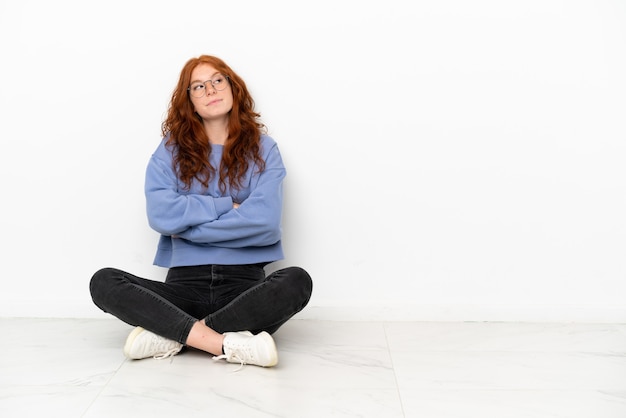 Ragazza rossa dell'adolescente che si siede sul pavimento isolata su fondo bianco che fa gesti di dubbi mentre solleva le spalle