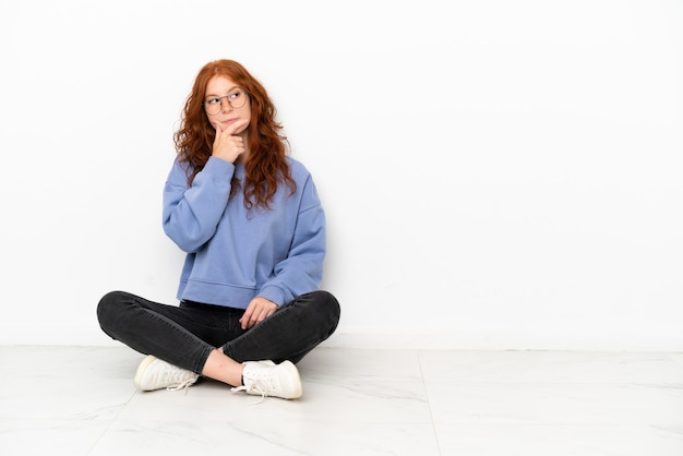 Ragazza rossa dell'adolescente che si siede sul pavimento isolata su fondo bianco che ha dubbi e con l'espressione confusa del viso