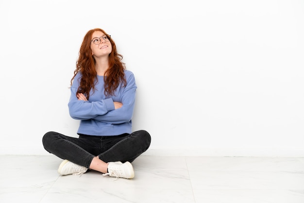 Ragazza rossa dell'adolescente che si siede sul pavimento isolata su fondo bianco che osserva in su mentre sorride