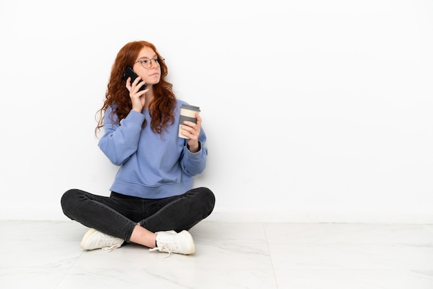 Ragazza rossa dell'adolescente che si siede sul pavimento isolata su fondo bianco che tiene caffè da portare via e un mobile