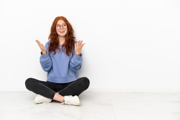 Ragazza rossa dell'adolescente che si siede sul pavimento isolato su fondo bianco con l'espressione facciale di sorpresa