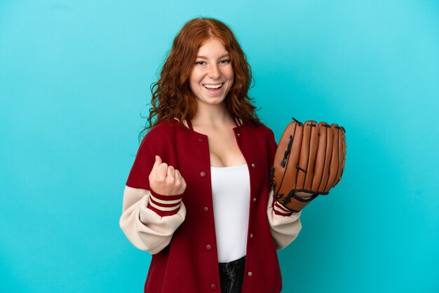 Ragazza rossa dell'adolescente con il guanto da baseball isolata su fondo blu che celebra una vittoria