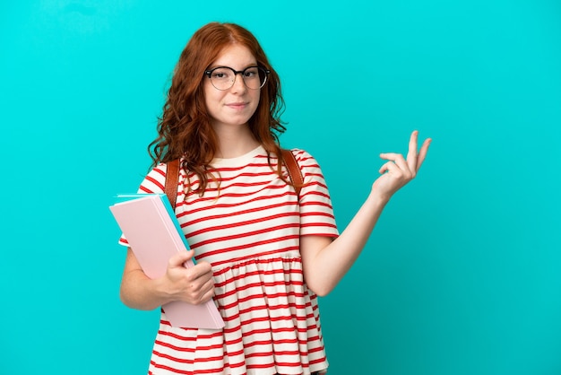 Ragazza rossa dell'adolescente dello studente isolata su fondo blu che estende le mani al lato per invitare a venire