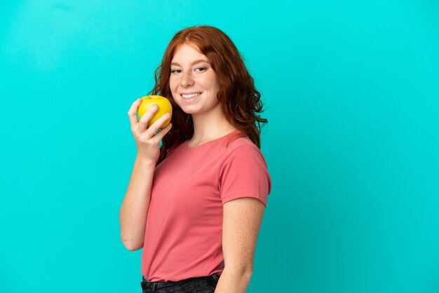 Ragazza rossa dell'adolescente isolata su fondo blu che mangia una mela
