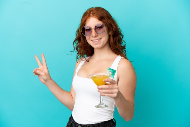 Ragazza rossa dell'adolescente isolata su fondo blu in costume da bagno e che tiene un cocktail