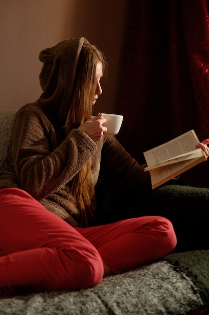 Ragazza rossa dello zenzero in divertente libro di lettura con cappuccio, bere caffè, sdraiato sul divano e sorridente