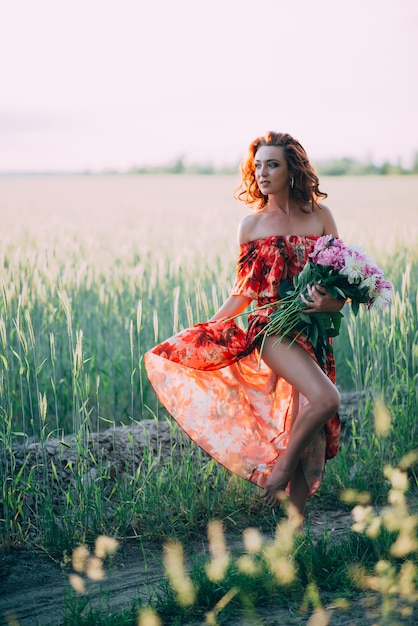 Ragazza rossa in abito rosso con bouquet di peonie danza gioiosa nel campo di grano in estate al tramonto.