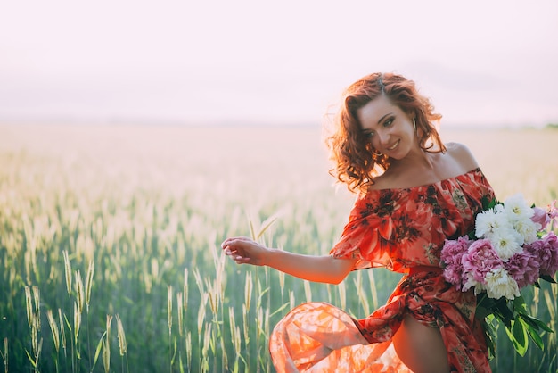 Ragazza rossa in abito rosso con bouquet di peonie danza gioiosa nel campo di grano in estate al tramonto.