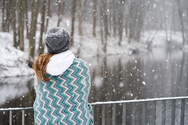 Ragazza rossa in cappello grigio, ricoperta di plaid blu modelli godendo nevicata in piedi sul retro sul ponte vicino al fiume e guardando il fiume