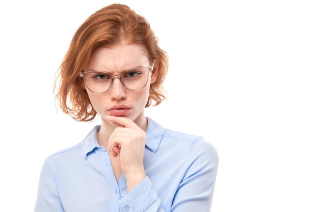 Ragazza rossa intelligente con gli occhiali e la camicia da lavoro che tiene il mento pensa che i dubbi prendano una decisione isolata su sfondo bianco per studio