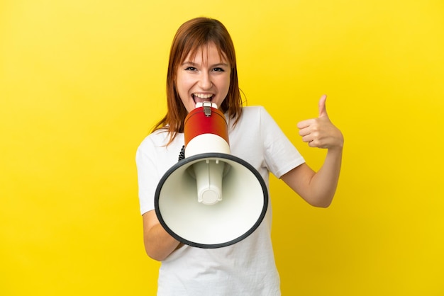 Ragazza rossa isolata su sfondo giallo che grida attraverso un megafono per annunciare qualcosa e con il pollice in su