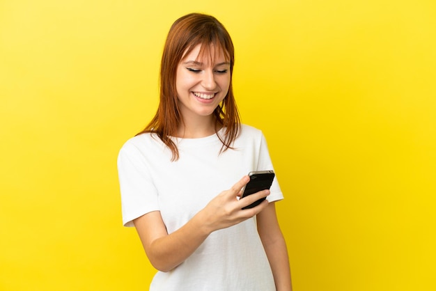 Ragazza rossa isolata su sfondo giallo che invia un messaggio o un'e-mail con il cellulare