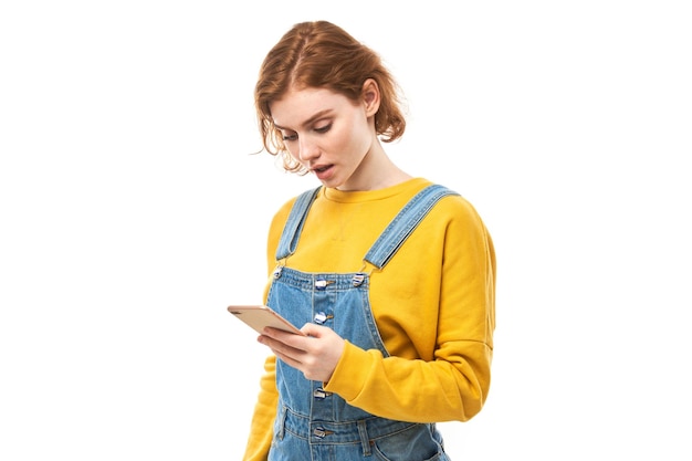 Ragazza rossa pensierosa che guarda lo schermo dello smartphone con la faccia sconvolta in abiti gialli isolati su sfondo bianco per studio
