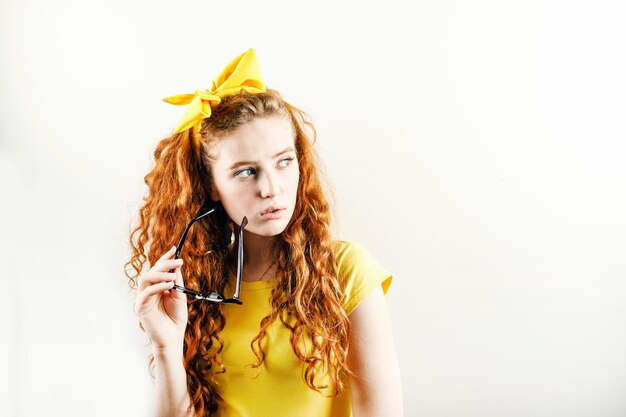 Ragazza rossa riccia pensierosa con un fiocco giallo sulla testa che indossa una t-shirt gialla con gli occhiali e distoglie lo sguardo