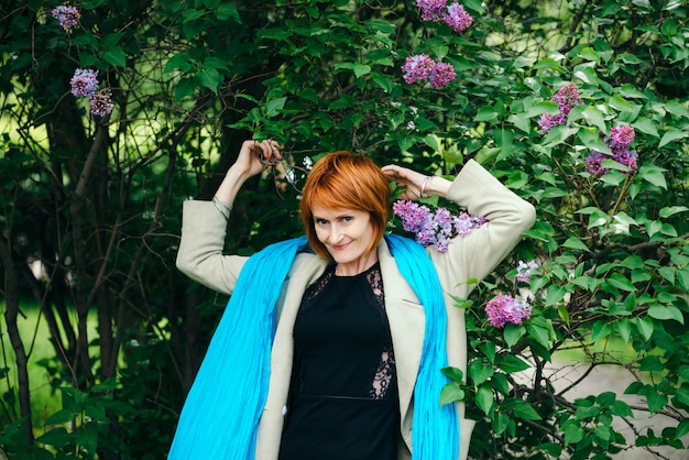 Ragazza rossa stupefacente dei capelli del hennè in vestito nero, cappotto luminoso e grande scialle blu che posano in cespugli del lillà di fioritura con lo spazio della copia.