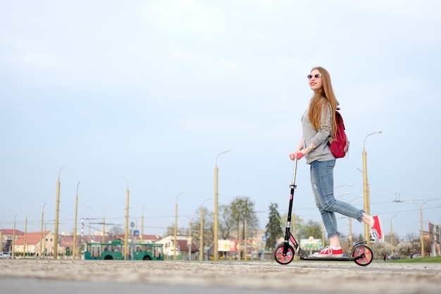 Ragazza rossa vestita di felpa con cappuccio grigia, blue jeans e scarpe da ginnastica rosse in sella a monopattino vicino a un edificio moderno