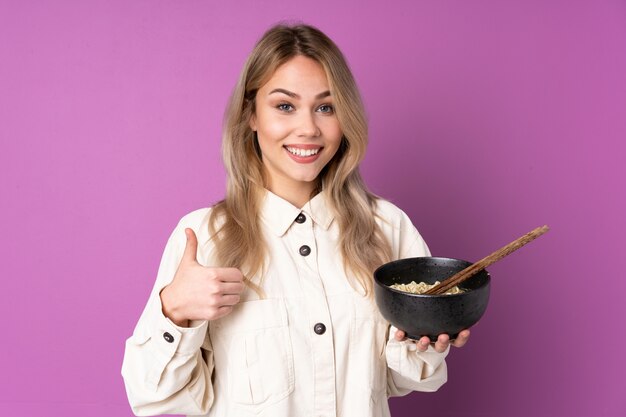 Ragazza russa adolescente sul muro viola con il pollice in alto perché è successo qualcosa di buono mentre si tiene una scodella di noodles con le bacchette