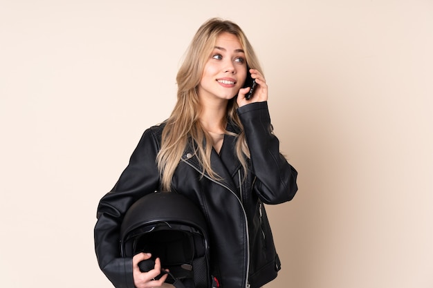 Ragazza russa con un casco da motociclista isolato sul muro beige mantenendo una conversazione con il telefono cellulare con qualcuno