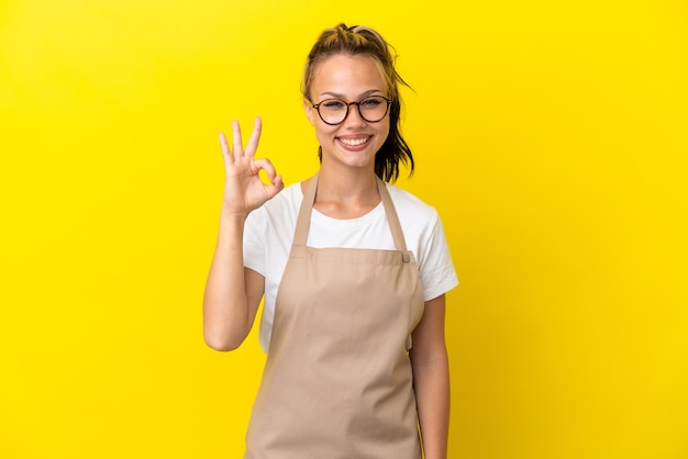 Ragazza russa del cameriere del ristorante isolata su fondo giallo che mostra il segno giusto con le dita