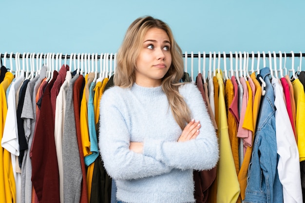 Ragazza russa dell'adolescente che compra dei vestiti isolati sulla parete blu che fa il gesto di dubbi mentre solleva le spalle