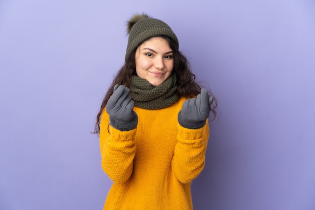 Ragazza russa dell'adolescente con il cappello di inverno isolato sulla parete viola che fa gesto dei soldi