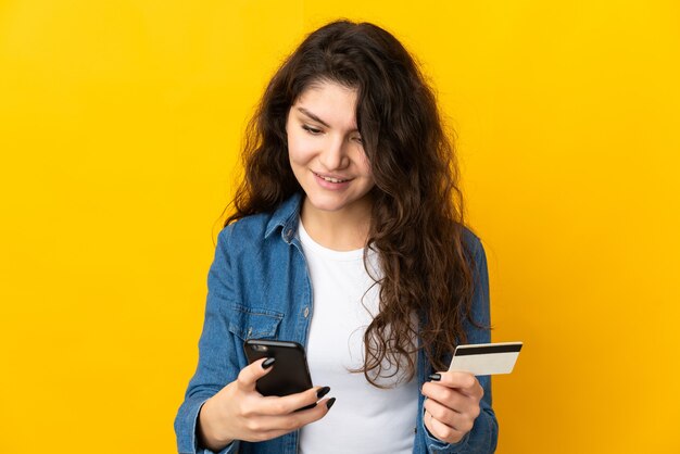 Ragazza russa dell'adolescente isolata su fondo giallo che compra con il cellulare con una carta di credito