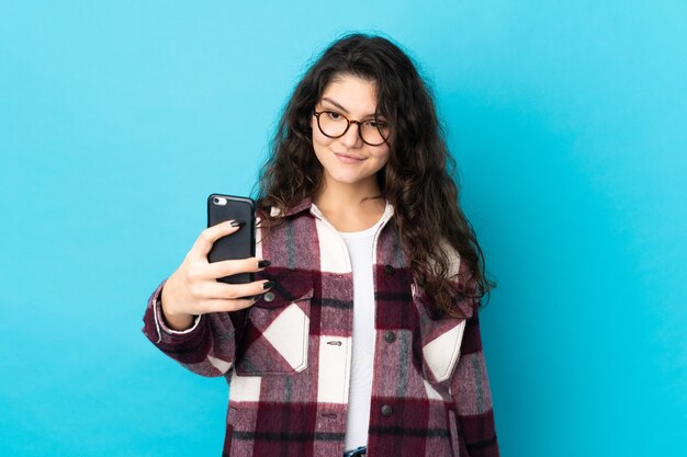 Ragazza russa dell'adolescente isolata sulla parete blu che fa un selfie