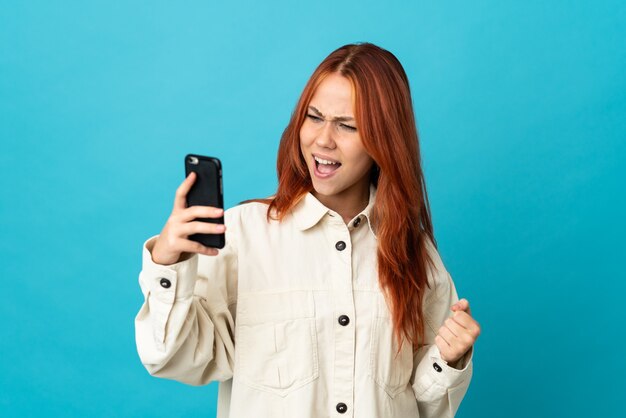 Ragazza russa dell'adolescente isolata sulla parete blu utilizzando il telefono cellulare e facendo il gesto di vittoria