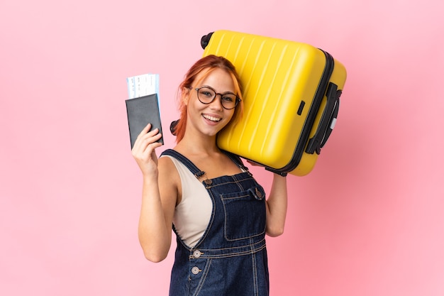 Ragazza russa dell'adolescente isolata sulla parete rosa in vacanza con la valigia e il passaporto