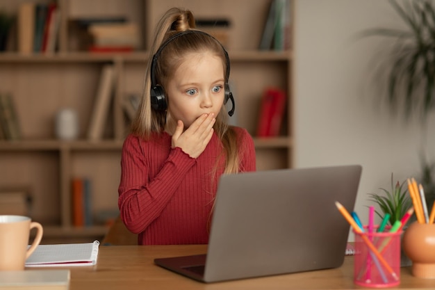 Ragazza scioccata del bambino che esamina il computer portatile che impara in linea all'interno