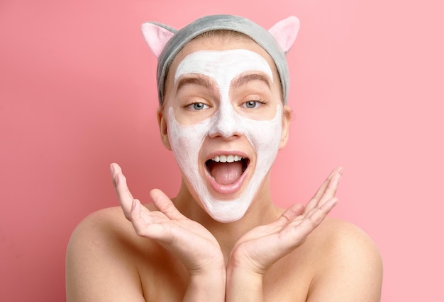Ragazza scioccata ottimista con maschera viso detergente cosmetica bianca e fascia per orecchie di gatto coreano alza i palmi delle mani, sorride gioiosamente, sorpresa su sfondo rosa