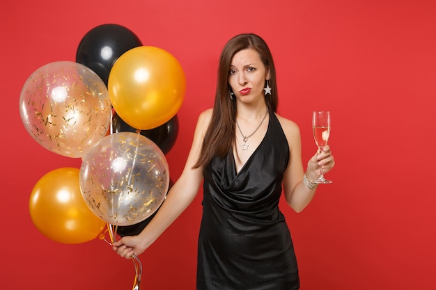 Ragazza sconvolta perplessa in abitino nero che celebra tenendo in mano un bicchiere di mongolfiere di champagne isolato su sfondo rosso. Giornata internazionale della donna Felice Anno Nuovo compleanno mockup festa concetto.