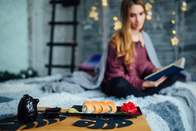 Ragazza seduta a letto con il libro accanto al pasto di sushi