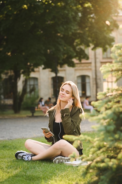 ragazza seduta sul prato nel parco universitario con libri e smartphone in mano ascoltando musica