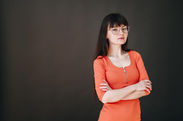 Ragazza seria con gli occhiali in un vestito lavorato a maglia ha piegato le mani e guarda la fotocamera su sfondo nero