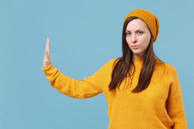 Ragazza seria giovane donna bruna in maglione giallo cappello in posa isolato su sfondo blu ritratto in studio. Concetto di stile di vita delle emozioni delle persone. Mock up spazio di copia. Mostra il gesto di arresto con il palmo a lato.