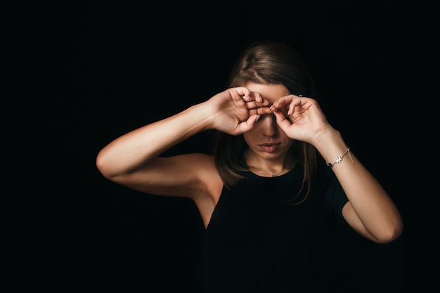 Ragazza seria su un muro nero. La ragazza si copre il viso con le mani. La donna ha seri problemi.