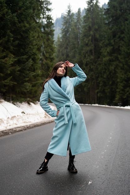 Ragazza sexy del brunette che tosa sulla strada sulla tabella delle montagne di inverno. Giovane donna affascinante che porta cappotto lungo blu alla moda. Concetto di moda, business e belle persone ..