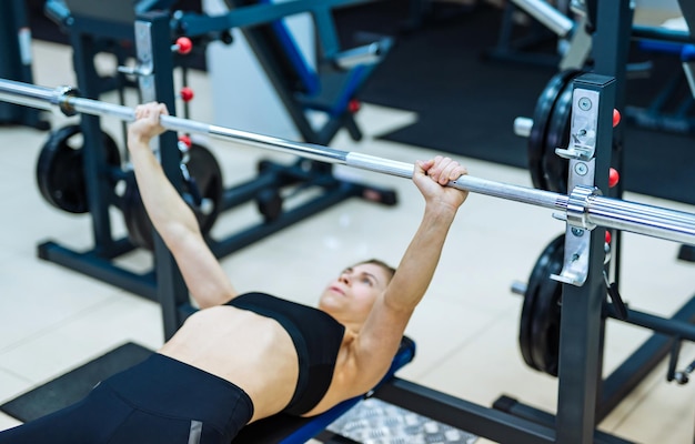 Ragazza sexy in forma sdraiata su un simulatore speciale e facendo esercizio di spinta con bilanciere che lavora sodo per muscoli forti al club sportivo Giovane sportiva in top nero e collant che si allena da sola