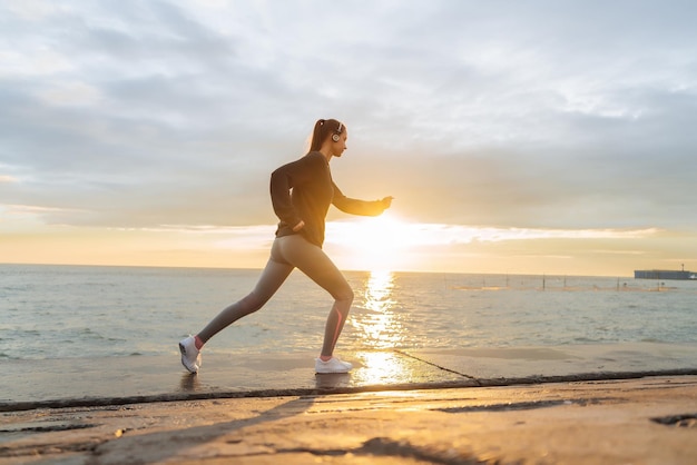 Ragazza sicura di sport che pareggia in riva al mare al tramonto ascoltando musica in cuffia