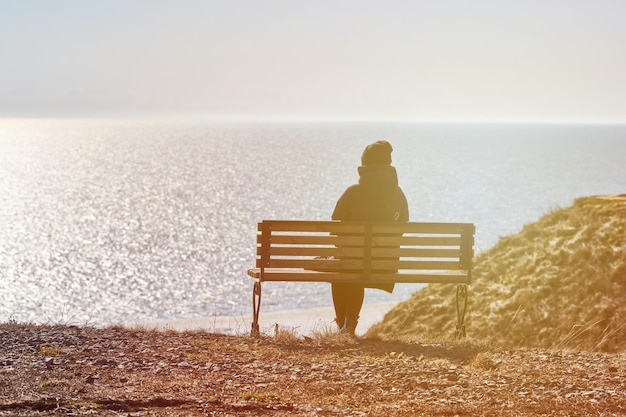 Ragazza single con una giacca nera e un cappello seduto su una panchina sulla scogliera di fronte al mare, posto tranquillo e tranquillo