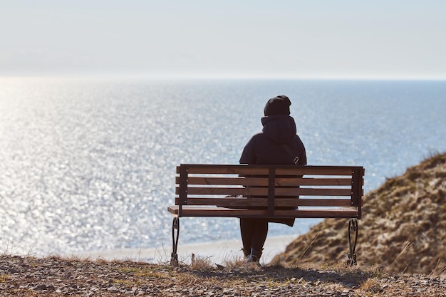 Ragazza single con una giacca nera e un cappello seduto su una panchina sulla scogliera di fronte al mare, posto tranquillo e tranquillo