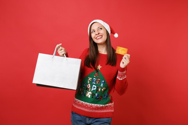 Ragazza sognante di Santa che guarda in alto, tiene in mano i sacchetti di pacchetti di carte di credito con gli acquisti dopo lo shopping isolato su sfondo rosso brillante. Felice Anno Nuovo 2019 celebrazione festa concetto. Mock up copia spazio.