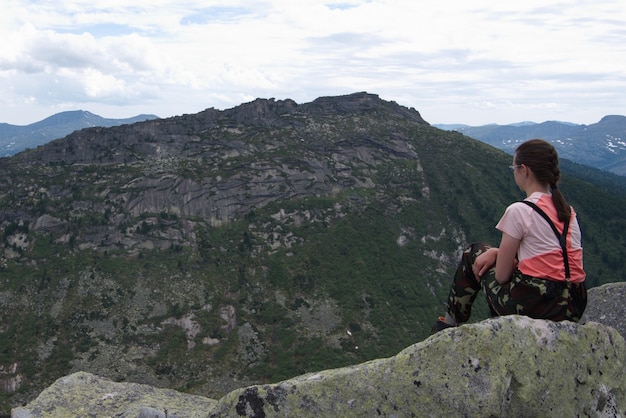 Ragazza solitaria giovane viaggiatore seduto su una pietra in cima a una montagna e guardando le alte montagne.